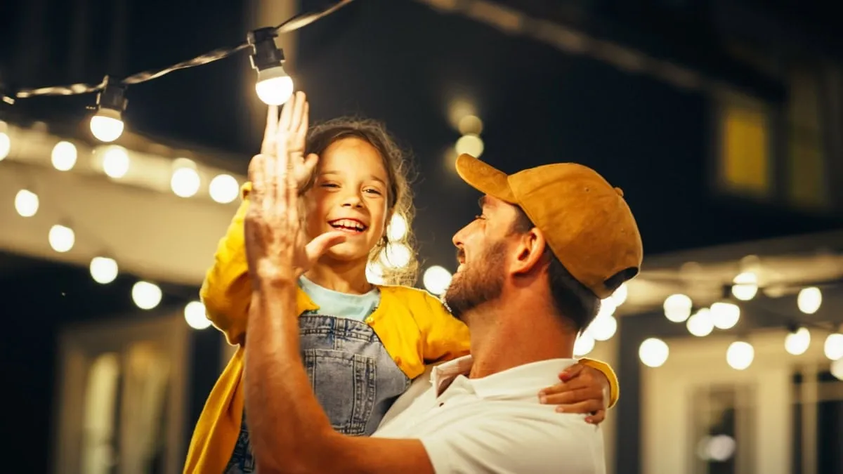 Father and daughter laughing together outdoors at night under string lights, both wearing yellow shirts.