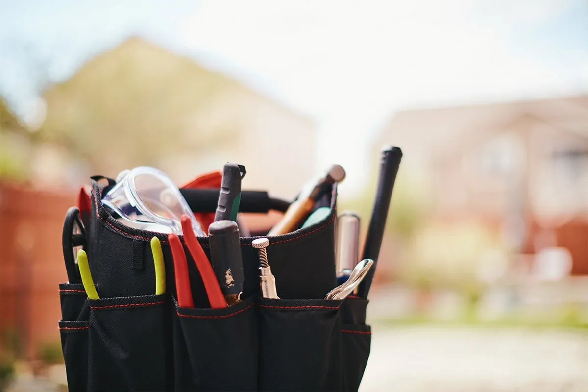 Black tool bag filled with various hand tools and safety glasses, residential houses blurred in background