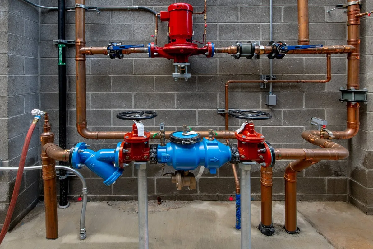 Commercial plumbing system with copper and PVC pipes, red and blue valves, and fire suppression equipment mounted on concrete block wall.