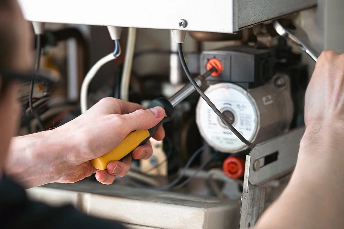 Technician holding yellow screwdriver while servicing HVAC unit interior with visible wiring and gauge meter.