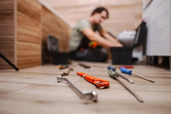 Work tools lying on bathroom floor while plumber is fixing a problem with toilet in the background.