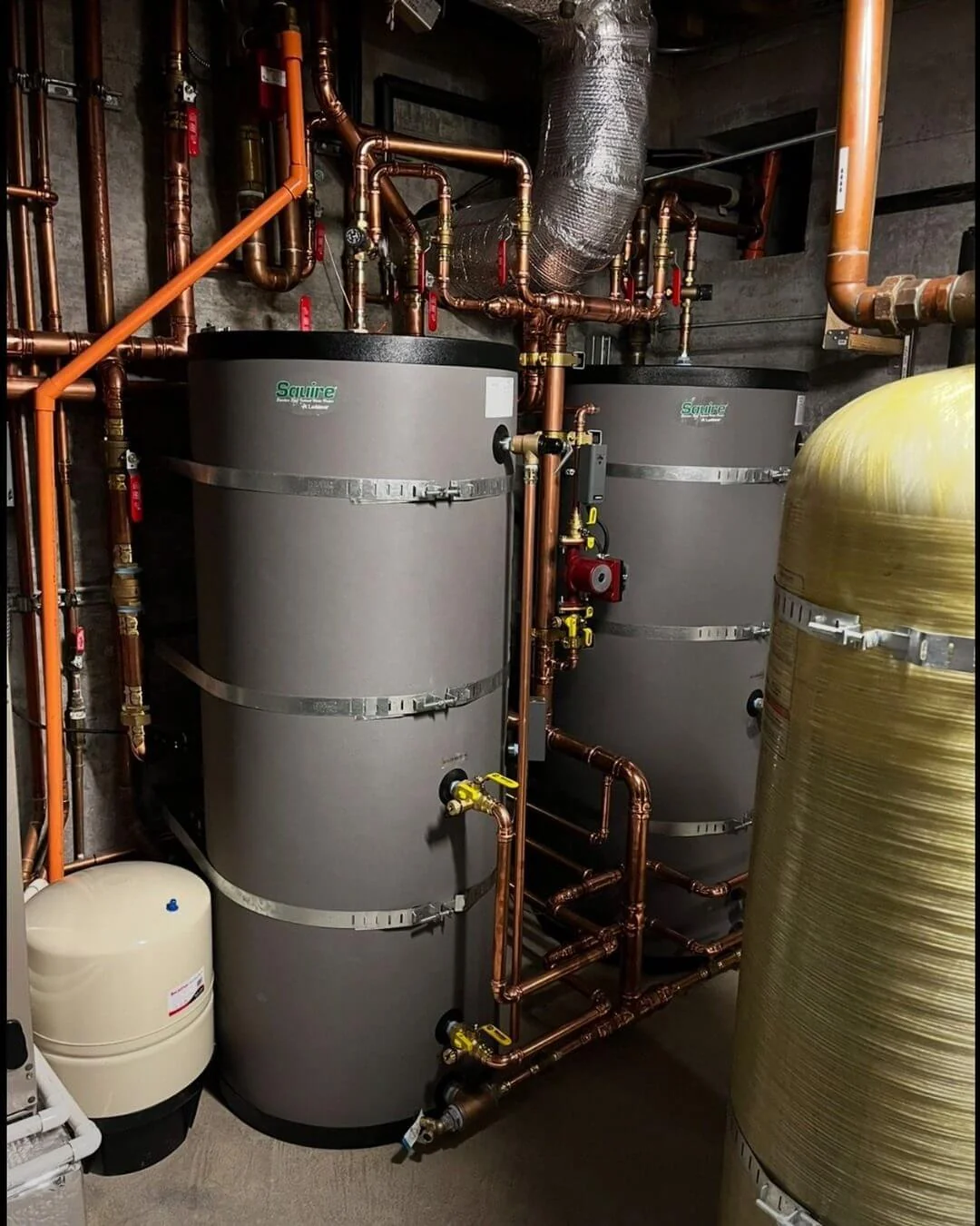 Two strapped storage water heaters joined by a copper manifold in a mechanical room, with an expansion tank and treatment vessel alongside