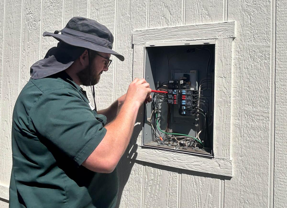 Ongaro electrician in green shirt and hat working on electrical panel mounted on white barn wall.